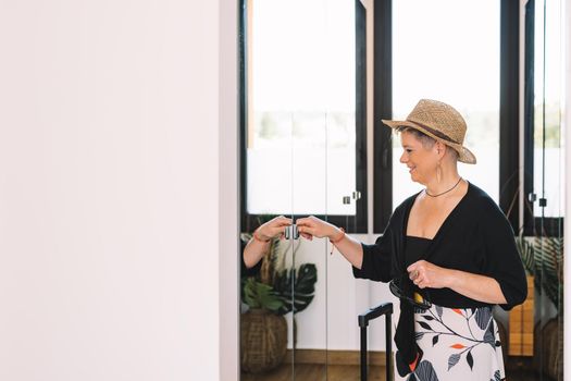 adventurous mature woman, arriving at the wardrobe of her summer holiday hotel room. businesswoman opening the wardrobe to leave her luggage. bedroom, natural light, dressing room with mirrors. copy space.