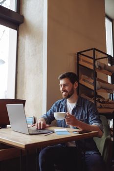 Happy caucasian young attractive man with cup of coffee sitting at laptop in cafe. Videocall concept