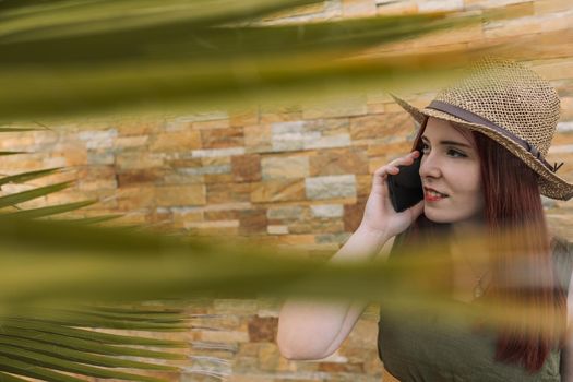 young red-haired girl, calling with her mobile phone to her summer holiday hotel. tourist in a tropical resort. business woman on a trip. outside, natural sunlight, palm trees and tropical flora.
