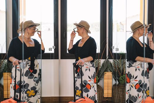 mature woman traveller, looking in the mirror while trying on her sunglasses for a walk along the coast on her summer holiday. bedroom, natural light, dressing room with mirrors.