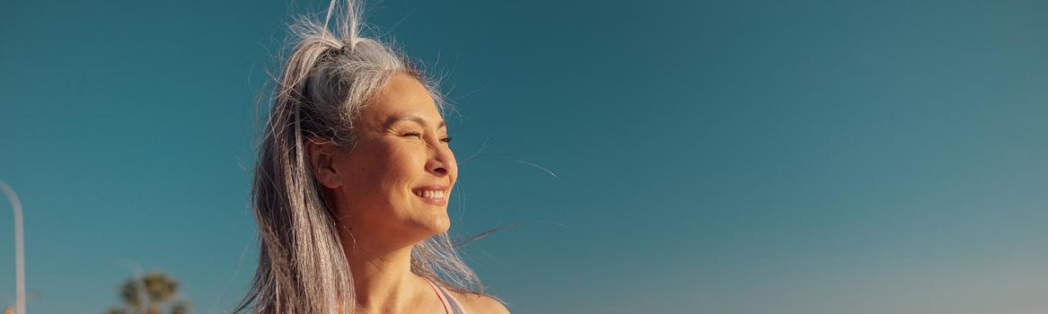 Sporty smiling female in sports top and leggings standing by the seashore and looking at the sun