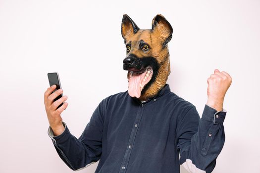 person with a dog mask looking at the phone and making winning gestures on white background, concept of success and technology
