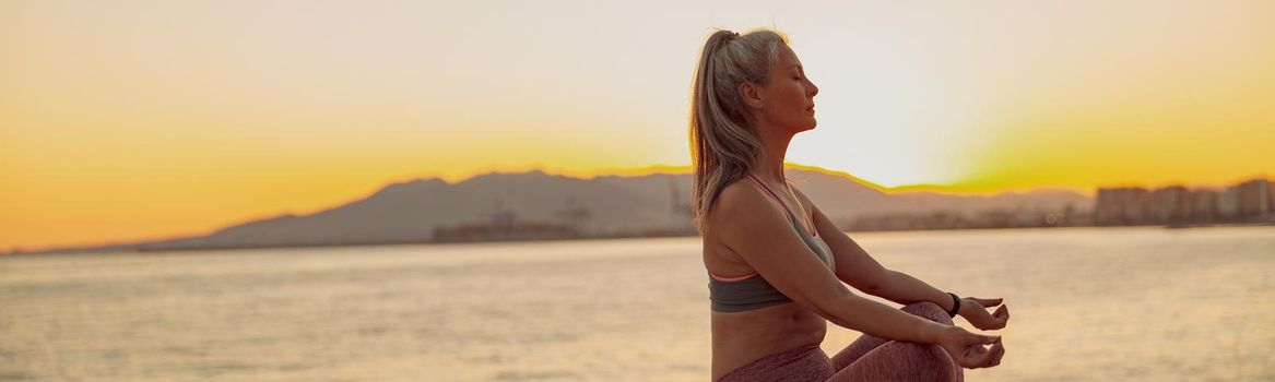 Side view of sporty lady practicing yoga, sitting on edge at seaside on background of sky at sunset and mountains