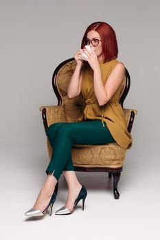Studio portrait of a beautiful luxury girl with chestnut hair. Red-haired girl is seated on a chair in a sexy suit and heels. Isolated on a gray background.