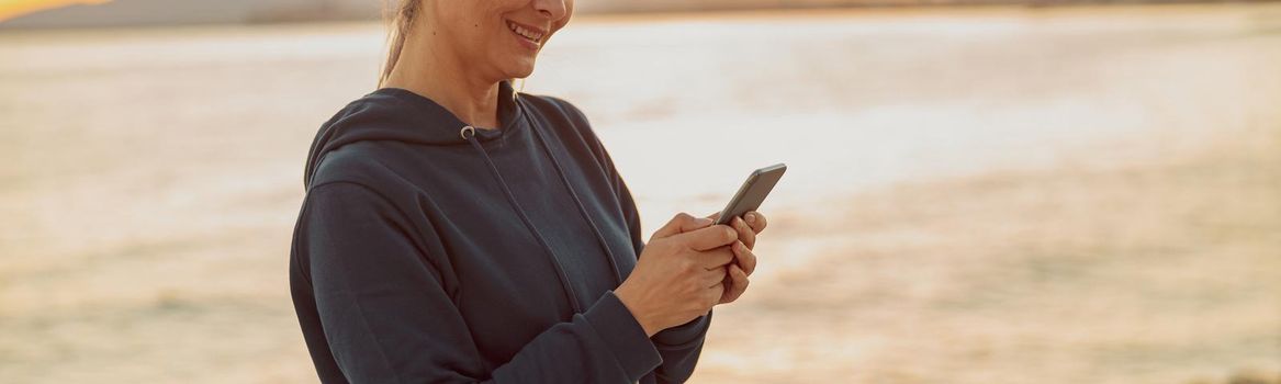 Charming sportswoman using smartphone and listening to music while standing on background of calm sea and sunset