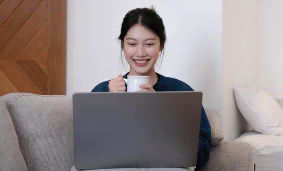 Asian Girl Working on Laptop Online, Using Internet and hold the cup. Sitting on Sofa at Home.