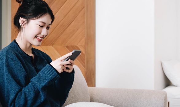 Smiling young asian woman using mobile phone while sitting on a couch at home with laptop computer.