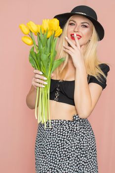 Summer fashion portrait blonde woman. Sexy look in black top and skirt, wearing hat. Red lips. Holding yellow tulips in her hands