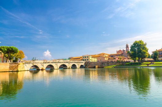 Famous stone arch Tiberius bridge Ponte di Tiberio Augustus over Marecchia river water, old buildings houses in historical touristic city centre Rimini with blue sky background, Emilia-Romagna, Italy