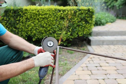 male hands in thick protective gloves cut an iron rod, sparks fly close-up. Cutting a metal rod with a burst of sparks. male repair work with electric tool in the garden.