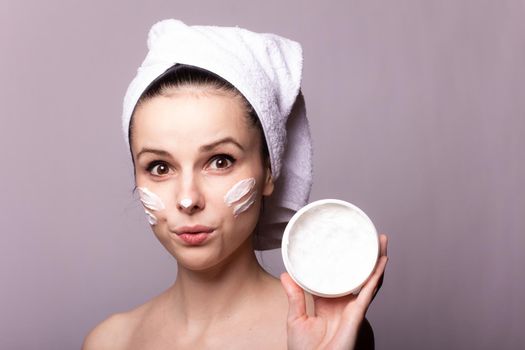 woman in a white towel on her head with cream on her face and a jar of cream in her hands, gray background. High quality photo