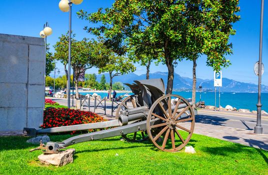 Cannon on green grass lawn in historical centre of Desenzano del Garda town with view of Garda Lake promenade embankment and mountain range, blue sky background, Lombardy, Northern Italy