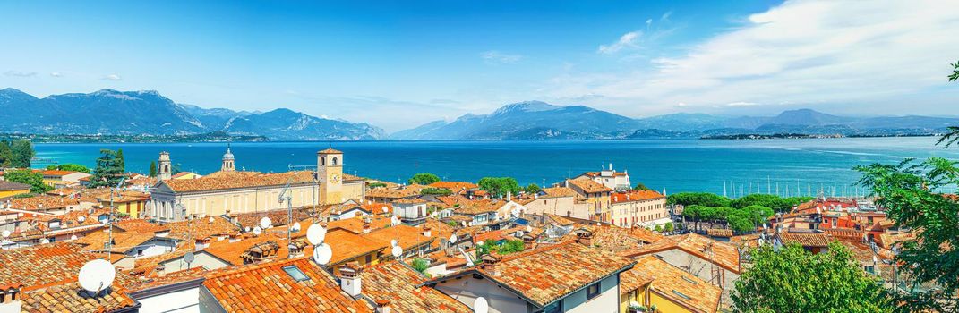 Panorama of Desenzano del Garda town with red tiled roof buildings, Garda Lake water, Monte Baldo mountain range, Sirmione peninsula, Lombardy, Northern Italy. Aerial panoramic view of Desenzano