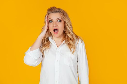 Confused European woman blonde frowning rounds her eyes, holding her hand on her head and looking at the camera with her mouth wide open, disappointed to hear bad news standing on a yellow background
