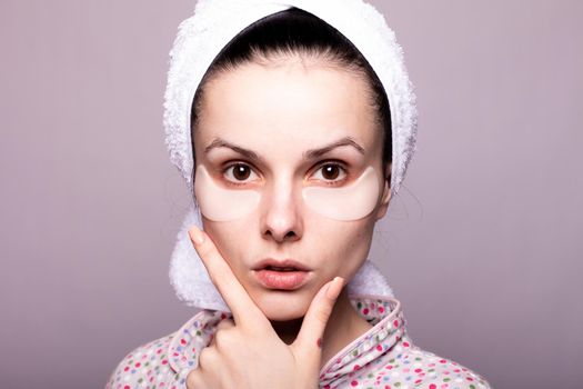 woman with cosmetic patches under her eyes, white towel on her head, gray background close-up. High quality photo