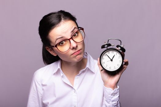 woman manager in a white shirt and glasses holds an alarm clock in her hand, gray studio background. High quality photo