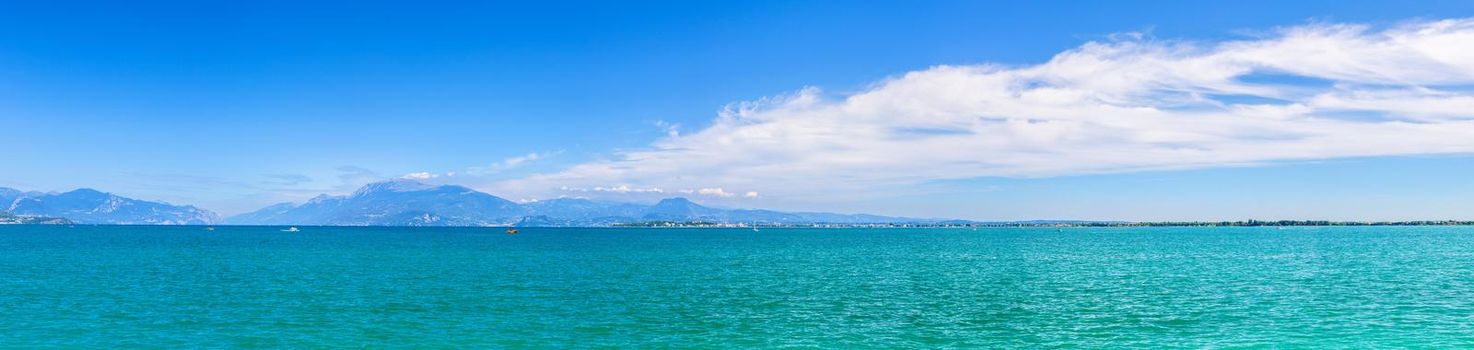 Panorama of Garda Lake azure turquoise water with view of Monte Baldo mountain range and Sirmione peninsula, blue sky white clouds background, Desenzano del Garda town, Lombardy, Northern Italy