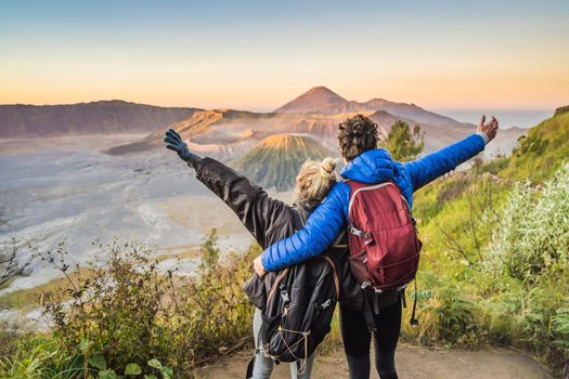 Young couple man and woman meet the sunrise at the Bromo Tengger Semeru National Park on the Java Island, Indonesia. They enjoy magnificent view on the Bromo or Gunung Bromo on Indonesian, Semeru and other volcanoes located inside of the Sea of Sand within the Tengger Caldera. One of the most famous volcanic objects in the world. Travel to Indonesia concept.