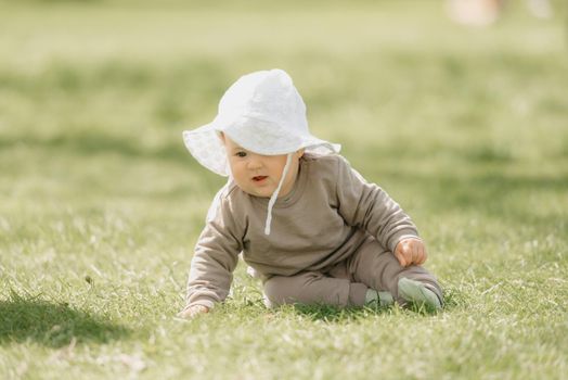 A young 7-month child in the panama hat is playing in the meadow. An infant girl is crawling on the grass.