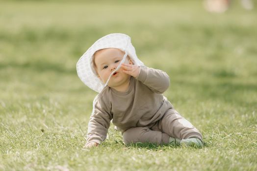 A young 7-month child in the panama hat is having fun in the meadow. An infant girl is crawling on the grass.
