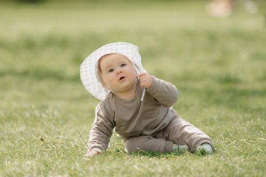 A young 7-month child in the panama hat is basking in the meadow. An infant girl is crawling on the grass.