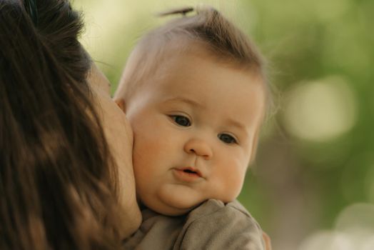 A close portrait of a 7-month girl who is been kissing by her mother in the park. A mom is holding in her arms her infant daughter in the woodland.