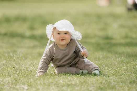 A smiling 7-month child in the panama hat is basking in the meadow. An infant girl is crawling on the grass.