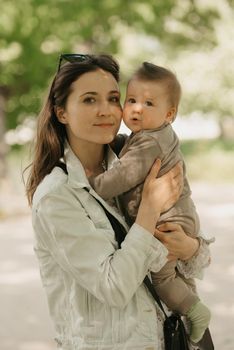 A young mother is holding in her arms her 7-month daughter in the park. A mom is happy with her infant daughter in the woodland.