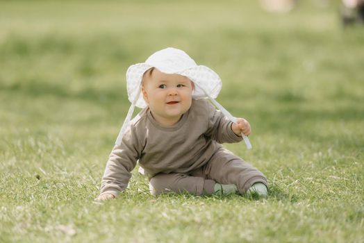 A 7-month child in the white panama hat is having fun in the meadow. An infant girl is crawling on the grass.