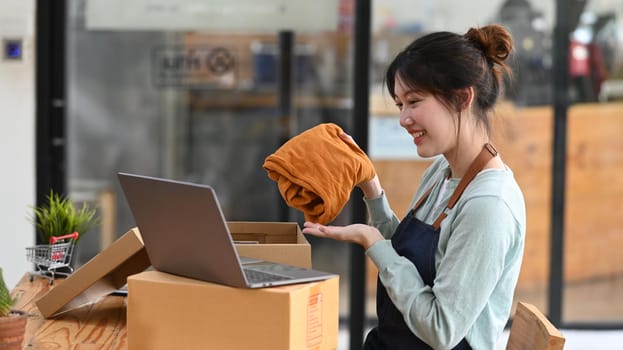 Shot of young female small business owner showing clothes while live streaming online on laptop computer.
