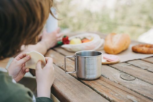 School boy kid child having a family picnic and looking at coffee, tea metallic mug that stands on a wooden table with bread and vegetables.