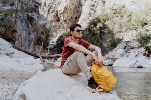 Man with yellow backpack sits on a riverside rock in the canyon with mountain cliffs in the background. Adult man taking a rest on a boulder near mountain river.