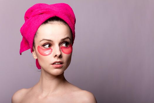 brunette woman with cosmetic pink patches under her eyes in a pink towel on her head, gray studio background, close-up portrait. High quality photo