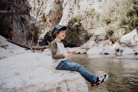 Caucasian school boy sits on a riverside rock in the canyon with mountain cliffs in the background. Kid child taking a rest on a boulder near mountain river bank.