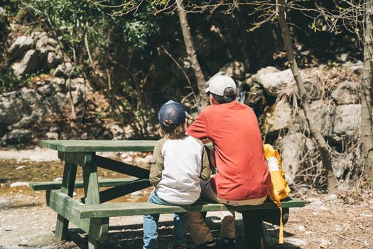 Rear view of tourists school boy and his dad sitting at the wooden table when taking a hike. Child boy and father wearing casual clothes and yellow backpack taking rest on a mountain hike.