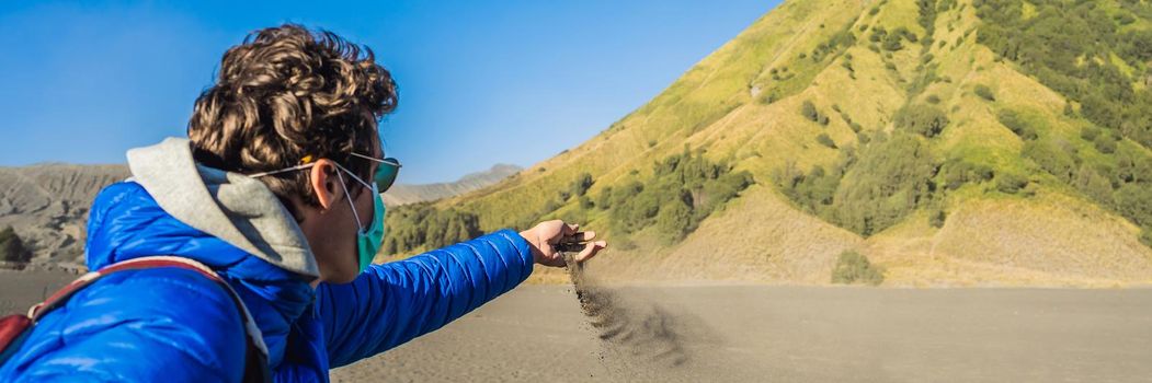 BANNER, LONG FORMAT Young man tourist touches volcanic sand in the Bromo Tengger Semeru National Park on the Java Island, Indonesia. He enjoys magnificent view on the Bromo or Gunung Bromo on Indonesian, Semeru and other volcanoes located inside of the Sea of Sand within the Tengger Caldera. One of the most famous volcanic objects in the world. Travel to Indonesia concept.