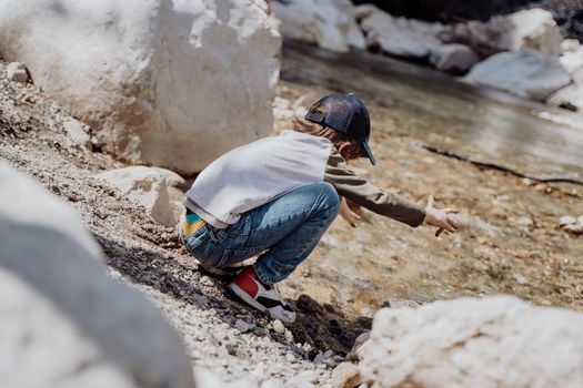 Caucasian school boy throwing rocks into the canyon river. Kid child playing with stones while sitting on a boulder near mountain river bank.