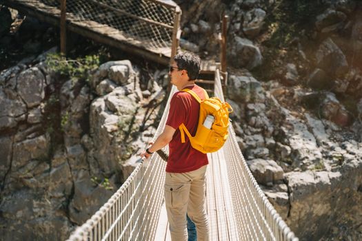 Father and son looking afar from the rope bridge in the mountains when hiking. Casually dressed tourists child school boy and his dad with yellow backpack crossing the canyon through rope bridge.