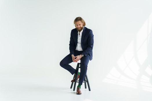 a handsome brutal bearded and long-haired man, a stylish businessman sitting on a chair with a white background. High quality photo