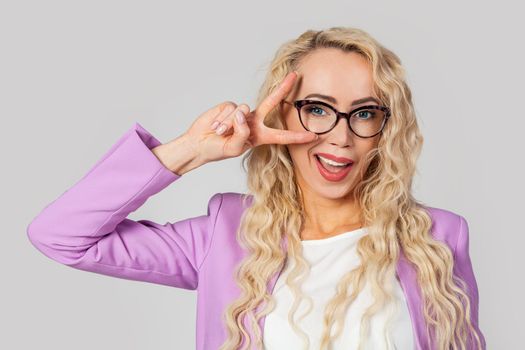 Portrait of a cheeky and cute glamorous blonde woman in glasses winking happily, showing a peace victory gesture as a fooling, optimistic emotion