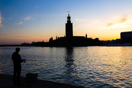 Silhouette of fisherman with fishing pole and Stockholm City Hall Stadshuset building Municipal Council on Kungsholmen Island at sunset, dusk, twilight with blue yellow orange sky background, Sweden
