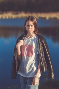 Portrait of a beautiful happy teenage girl in leather jacket outdoors in spring. A girl poses on lakes shore.