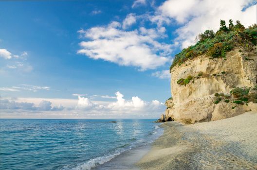 Rock with greenery near sandy beach shore coastline of Tyrrhenian Sea with blue sky and white clouds, Tropea town, Vibo Valentia, Calabria, Southern Italy