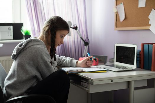 At home in her room, the teenager sits at her desk and solves assignments from school. A diligent student. Remote learning at home.