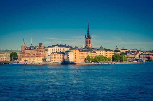 Riddarholmen island district with Riddarholm Church spires and typical sweden colorful gothic buildings, boat ship moored on Lake Malaren water from Kungsholmen with blue clear sky, Stockholm, Sweden