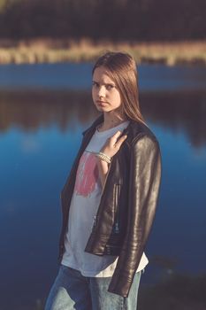 Portrait of a beautiful happy teenage girl in leather jacket outdoors in spring. A girl poses on lakes shore.