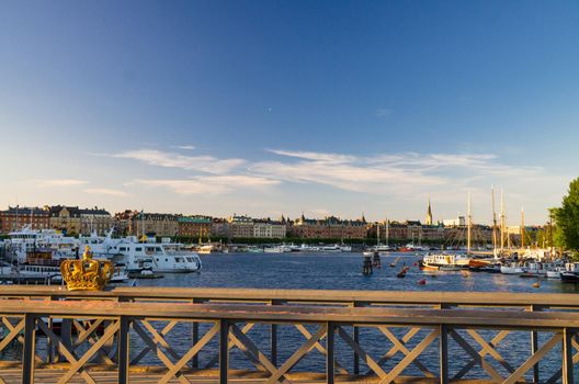 Golden Gilded Crown on Skeppsholmsbron bridge wooden railing with view of Ostermalm district with traditional typical buildings and yachts, ships, boats on Lake Malaren water, Stockholm, Sweden