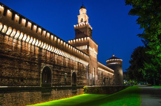 Old medieval Sforza Castle Castello Sforzesco lightning facade, walls, tower La torre del Filarete with lights, trees at sunset, dusk, twilight, evening, blue sky background, Milan, Lombardy, Italy