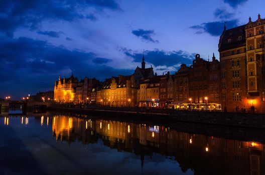 Typical buildings and Green gate Zielona Brama at Long Bridge embankment promenade reflection in Motlawa river water in old historical town centre, street lights dark blue sky evening, Gdansk, Poland