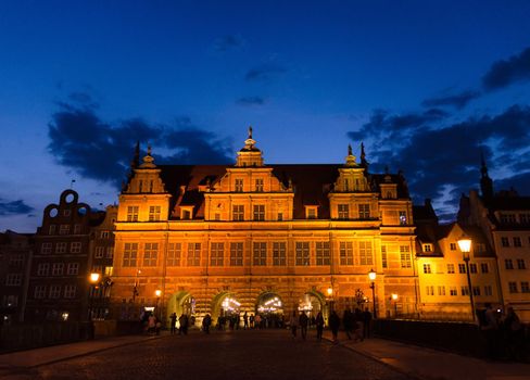 Green gate Zielona Brama building and street lamps lights on bridge over Motlawa river and dark blue sky background in the evening at night, sunset, dusk, twilight, Gdansk, Pomerania, Poland
